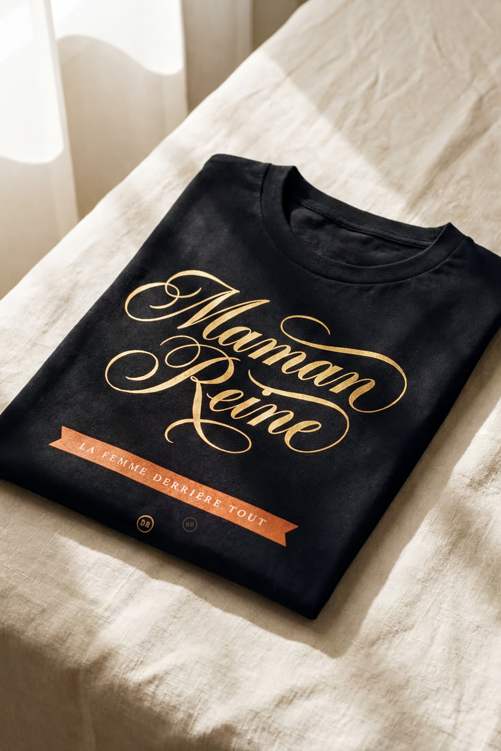 Black Maman Reine tee folded flat on natural linen, gold calligraphy and terracotta banner, Essentials gift line by Bana Bilaka