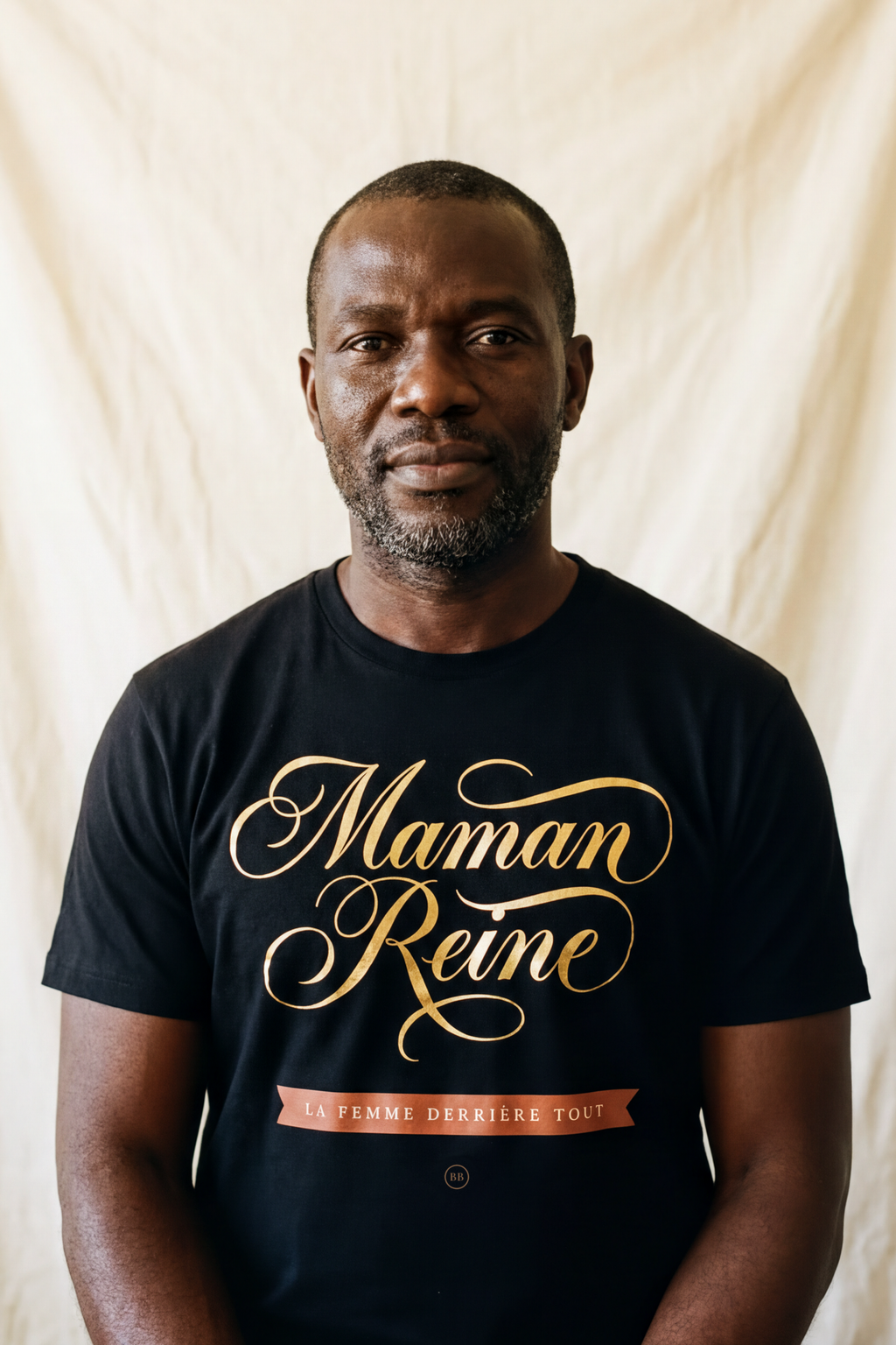 Man in his forties with a greying beard wearing the black Maman Reine calligraphy tee, banner reading La Femme Derrière Tout, Essentials gift line by Bana Bilaka