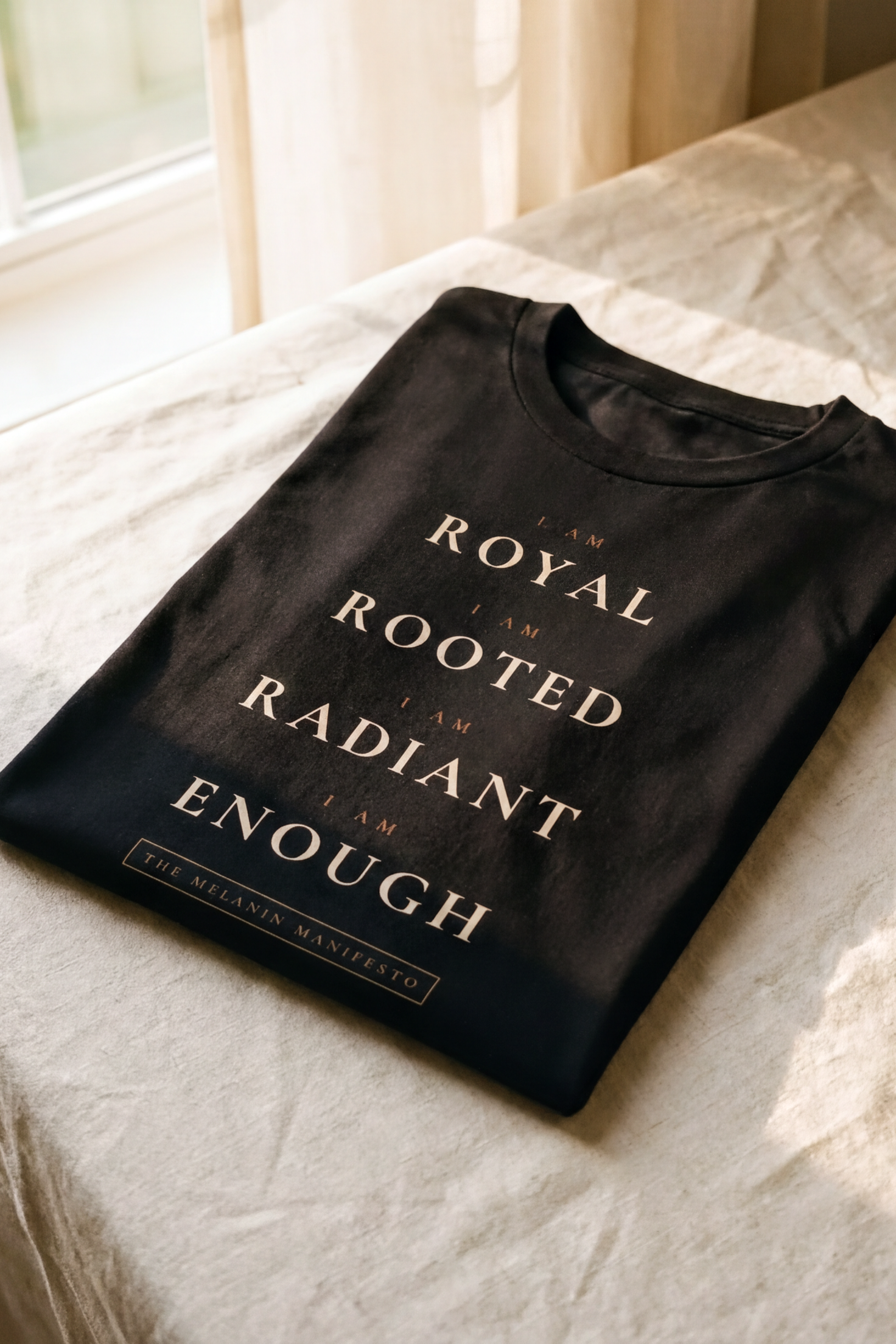 Black Melanin Manifesto V2 tee folded flat on natural linen, four-line litany I Am Royal Rooted Radiant Enough, Essentials collection by Bana Bilaka