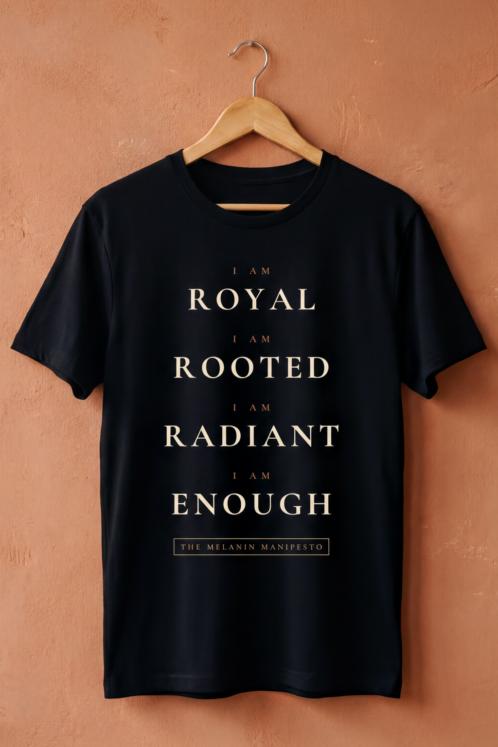 Black Melanin Manifesto V2 tee on a wooden hanger against a terracotta wall, Essentials collection by Bana Bilaka