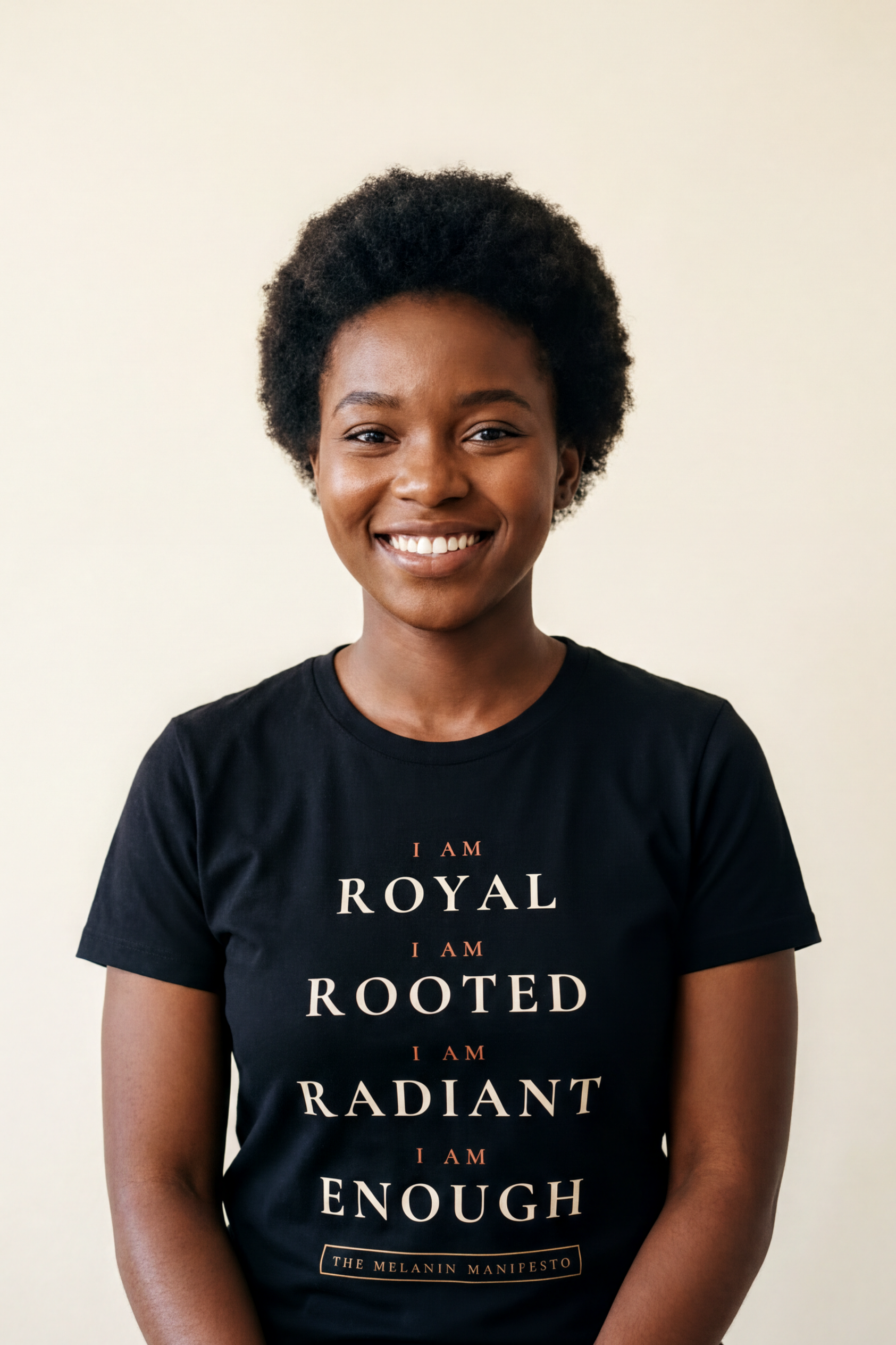 Smiling young woman with a short natural afro wearing the black Melanin Manifesto V2 tee, I Am litany in ivory and terracotta, Essentials collection by Bana Bilaka