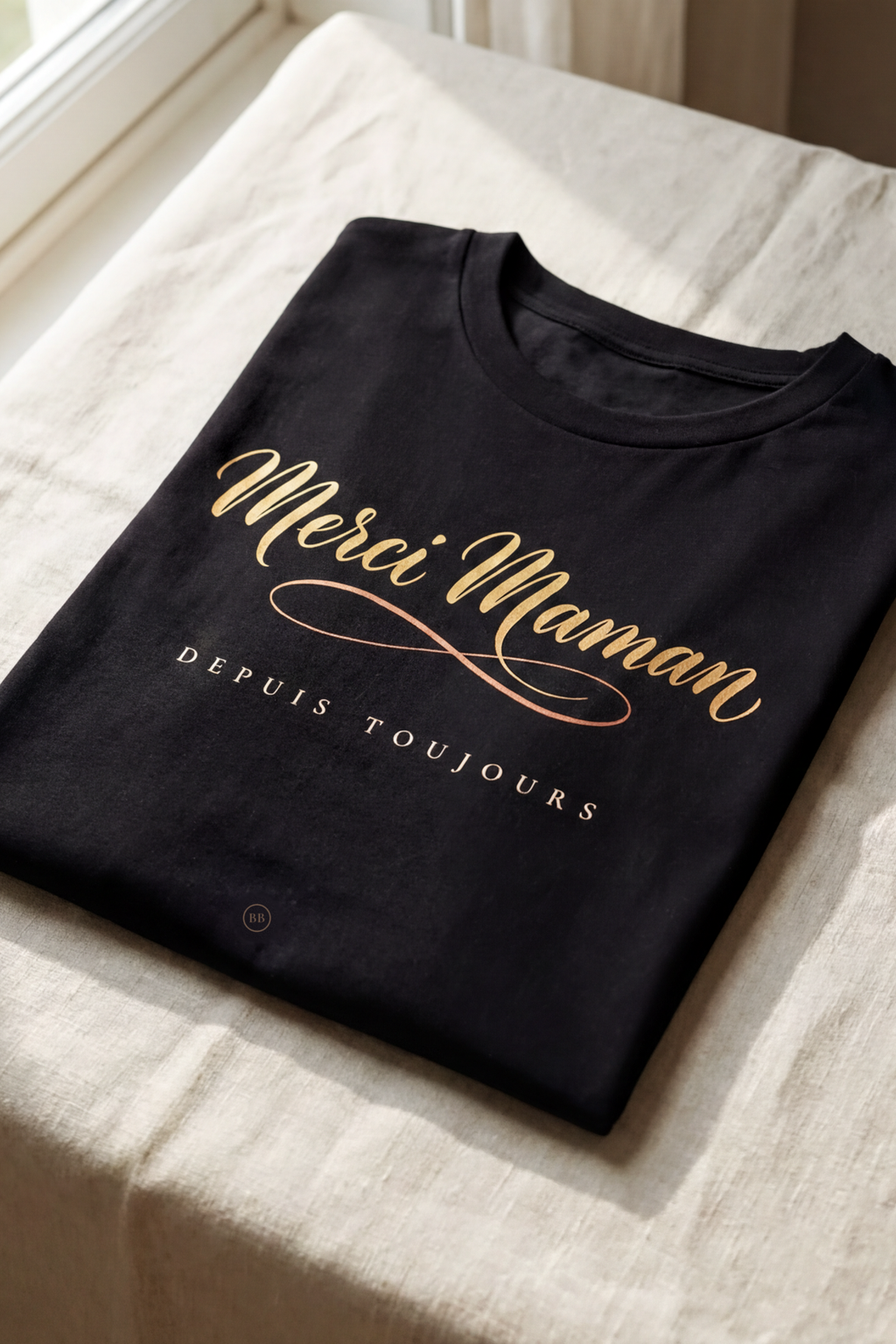 Black Merci Maman tee folded flat on natural linen, gold French calligraphy with the line Depuis Toujours, Essentials gift line by Bana Bilaka