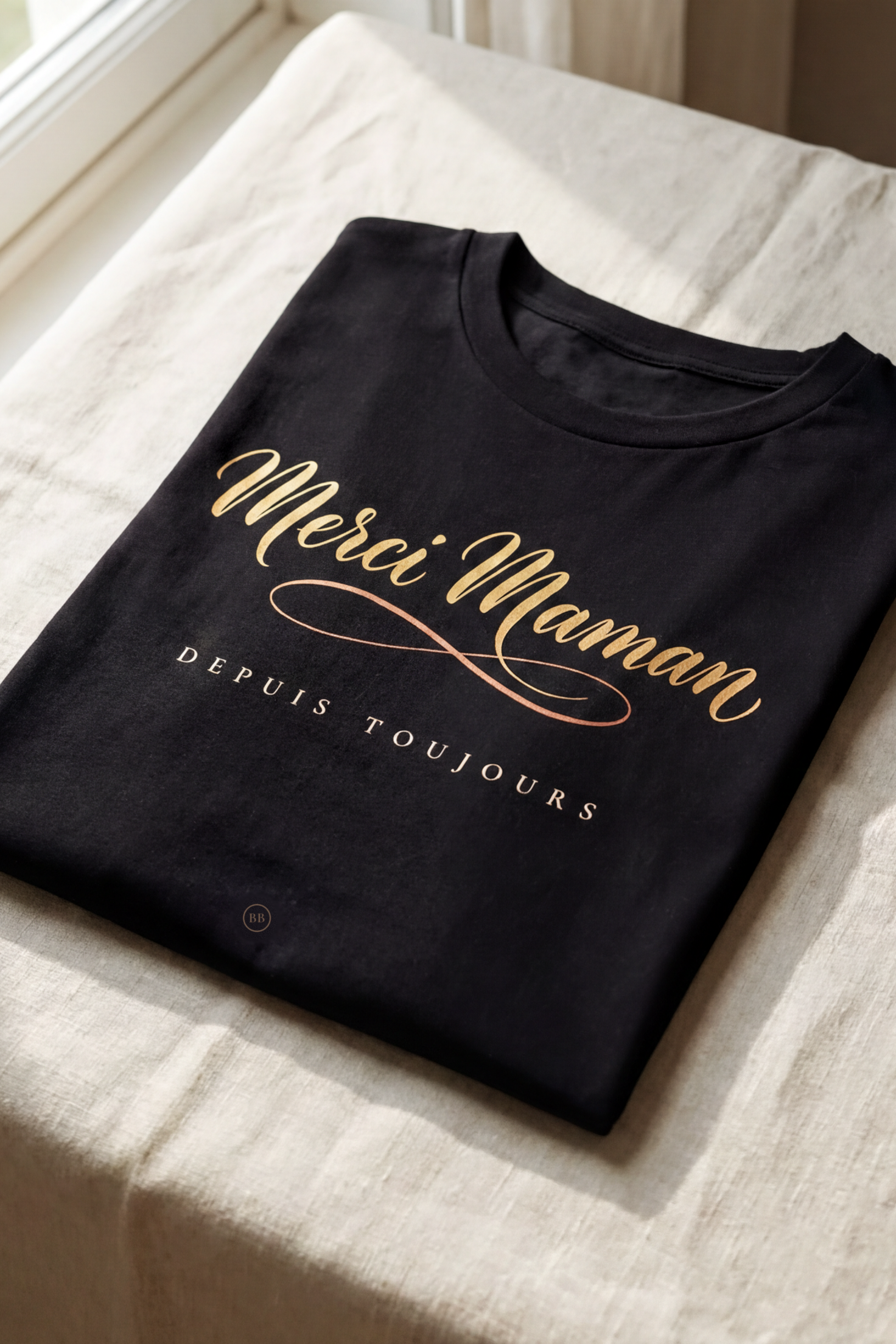 Black Merci Maman tee folded flat on natural linen, gold French calligraphy with the line Depuis Toujours, Essentials gift line by Bana Bilaka