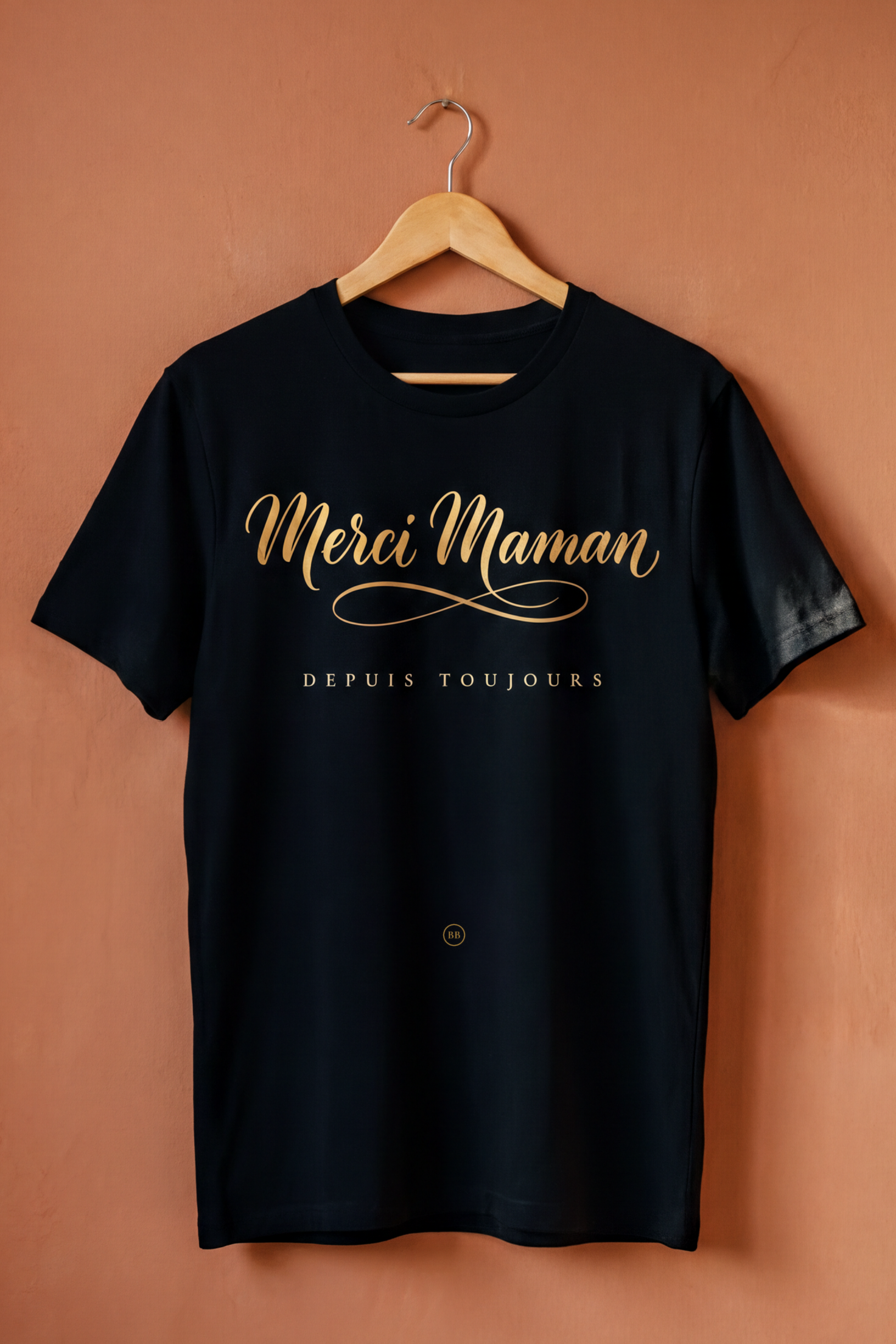 Black Merci Maman tee on a wooden hanger against a terracotta wall, Essentials gift line by Bana Bilaka