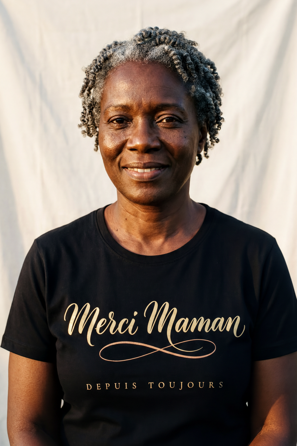 Woman in her fifties with natural silver coils wearing the black Merci Maman tee, gold French calligraphy with terracotta flourish, Essentials gift line by Bana Bilaka