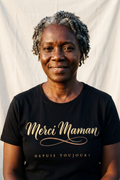 Woman in her fifties with natural silver coils wearing the black Merci Maman tee, gold French calligraphy with terracotta flourish, Essentials gift line by Bana Bilaka