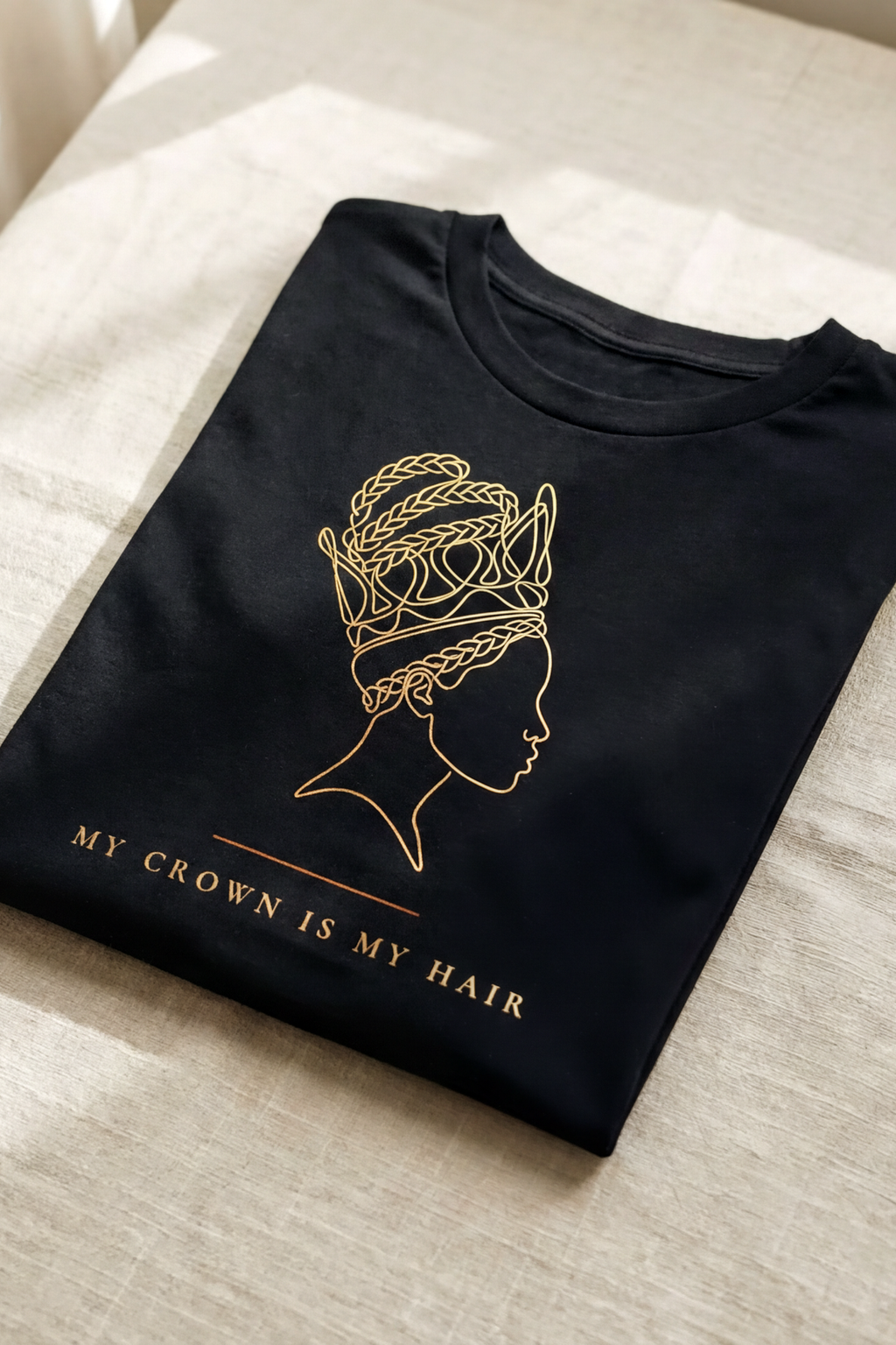 Black My Crown Is My Hair tee folded flat on natural linen, single-line gold silhouette with braided crown hair, Essentials collection by Bana Bilaka