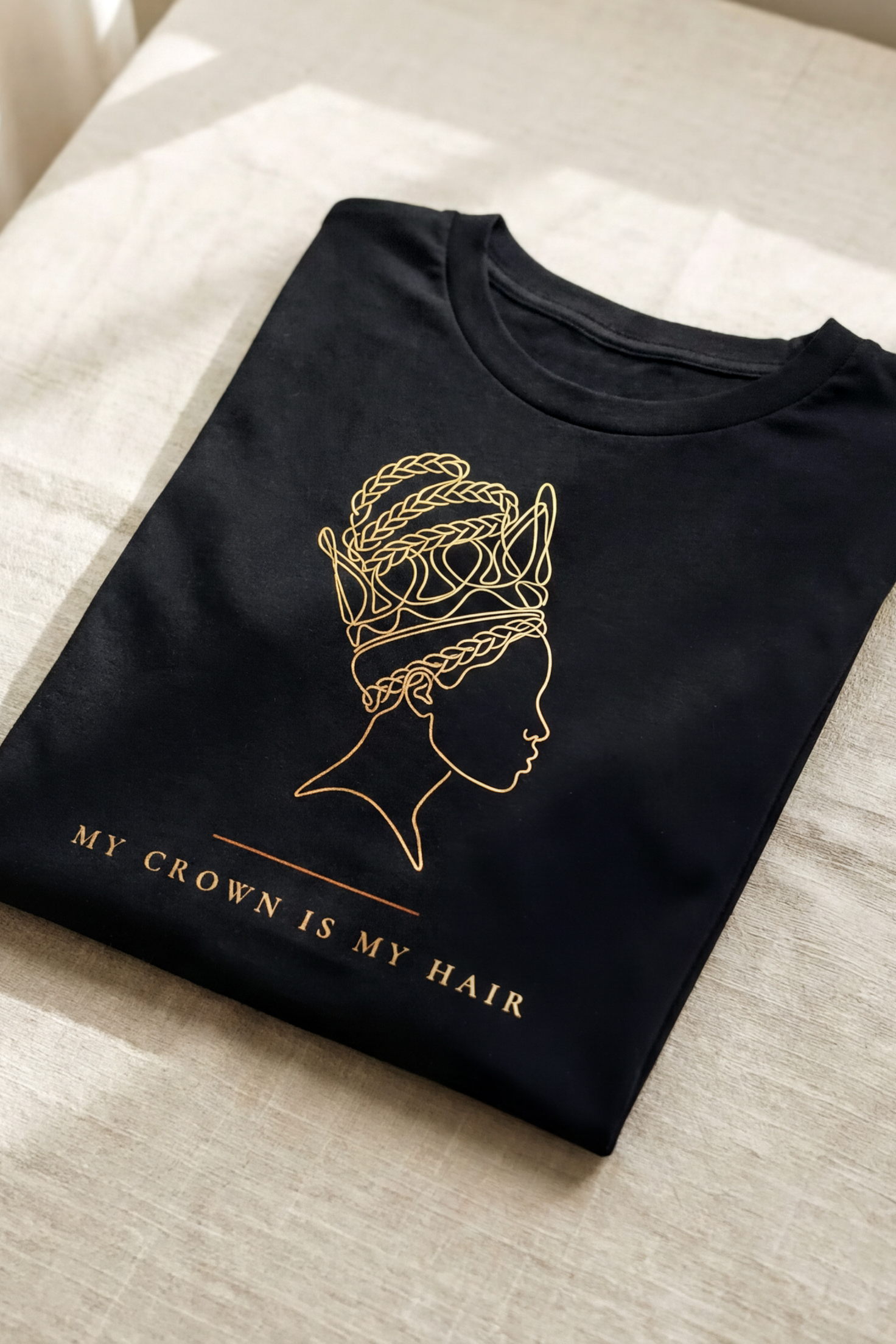 Black My Crown Is My Hair tee folded flat on natural linen, single-line gold silhouette with braided crown hair, Essentials collection by Bana Bilaka