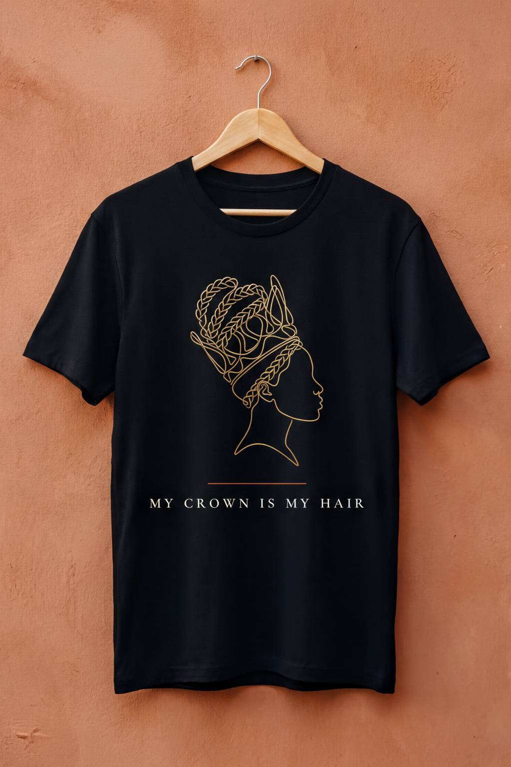 Black My Crown Is My Hair tee on a wooden hanger against a terracotta wall, Essentials collection by Bana Bilaka