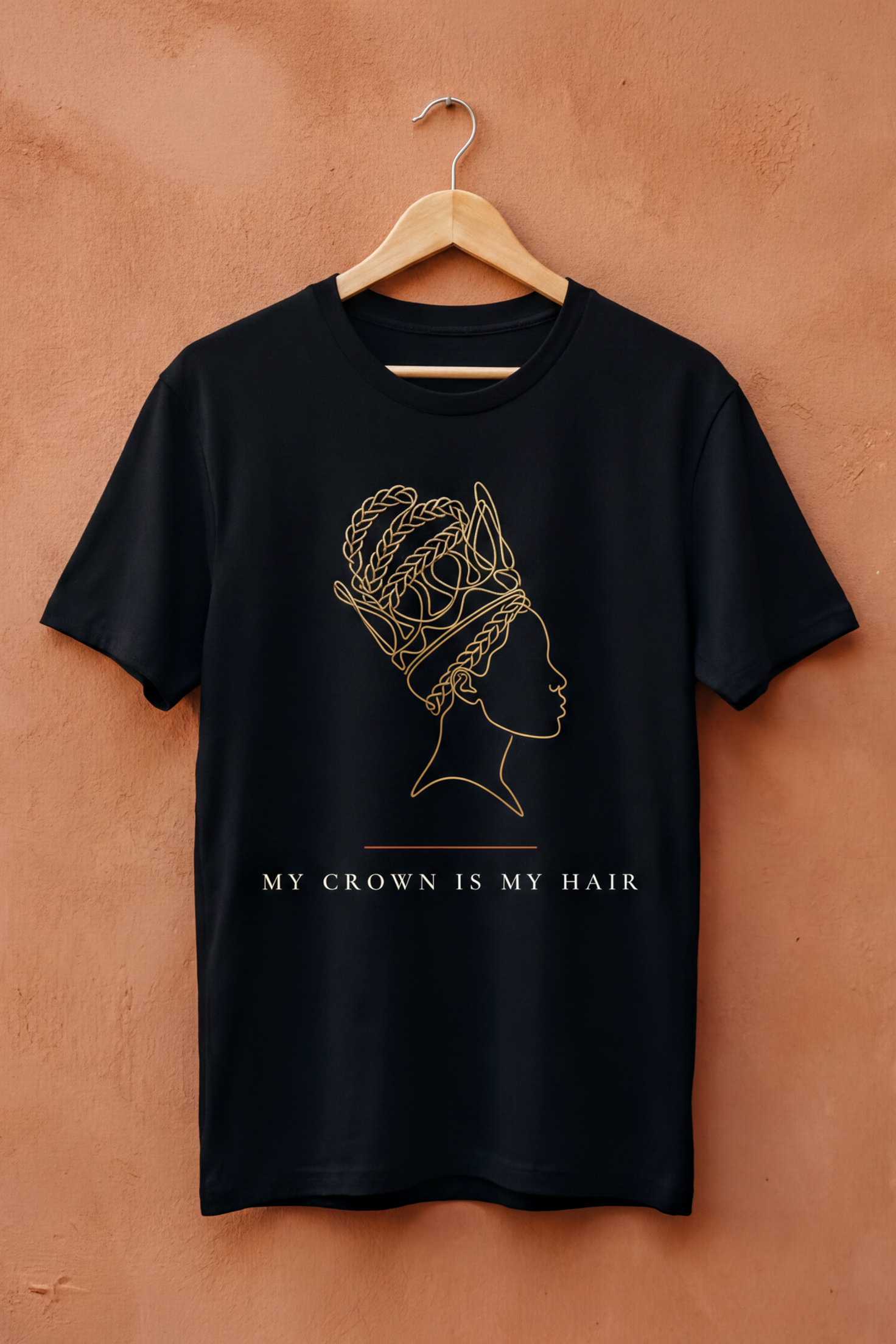 Black My Crown Is My Hair tee on a wooden hanger against a terracotta wall, Essentials collection by Bana Bilaka