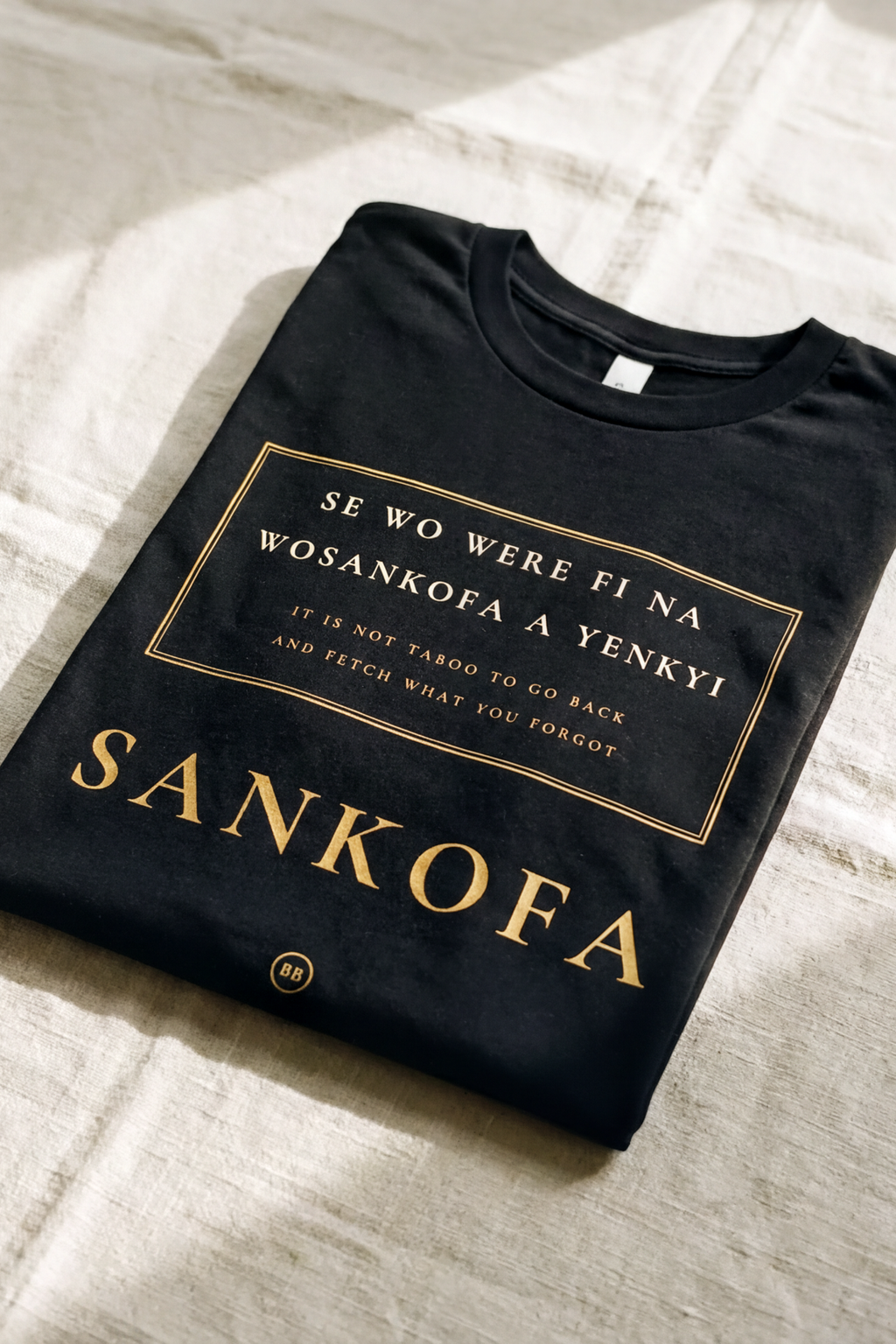 Black Sankofa V2 tee folded flat on natural linen, double gold rule cartouche with the full Akan proverb and the word Sankofa, Essentials collection by Bana Bilaka