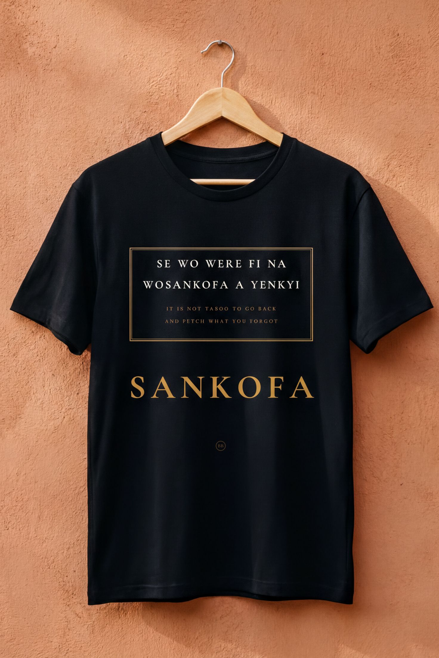 Black Sankofa V2 tee on a wooden hanger against a terracotta wall, Essentials collection by Bana Bilaka