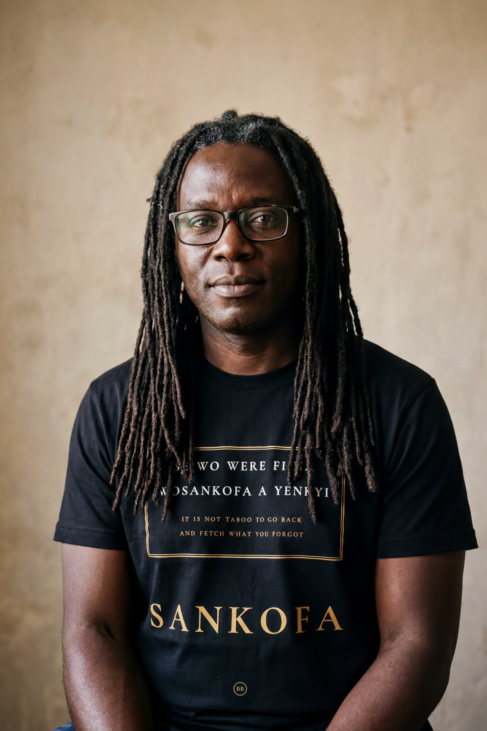 Man with long mature natural locs and glasses wearing the black Sankofa V2 tee by a window, Akan proverb cartouche in gold, Essentials collection by Bana Bilaka