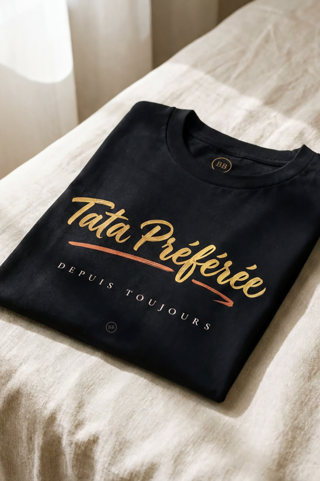 Black Tata Préférée tee folded flat on natural linen, gold calligraphy with terracotta brush stroke and Depuis Toujours line, Essentials gift line by Bana Bilaka