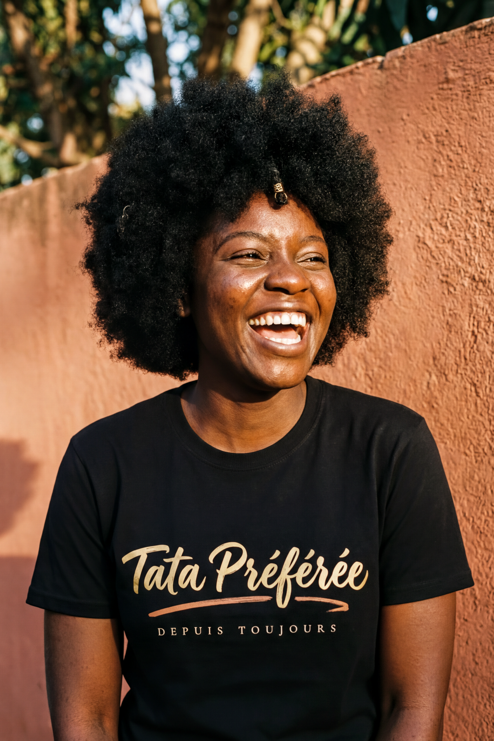 Laughing woman with a natural afro and gold hair ring wearing the black Tata Préférée tee, joyful gold calligraphy, Essentials gift line by Bana Bilaka
