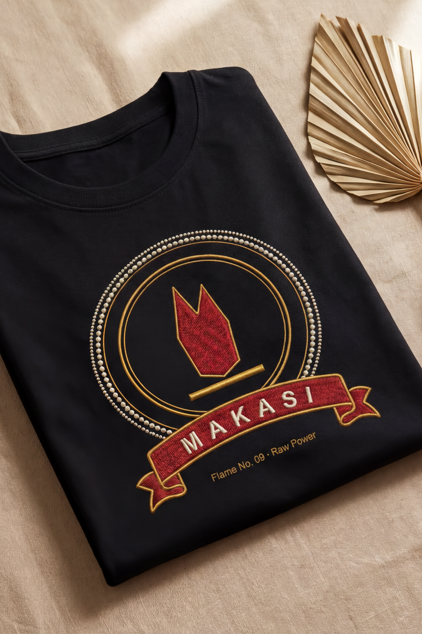 Black Makasi tee folded flat on natural linen, ruby raw power medallion, The Twelve Flames by Bana Bilaka