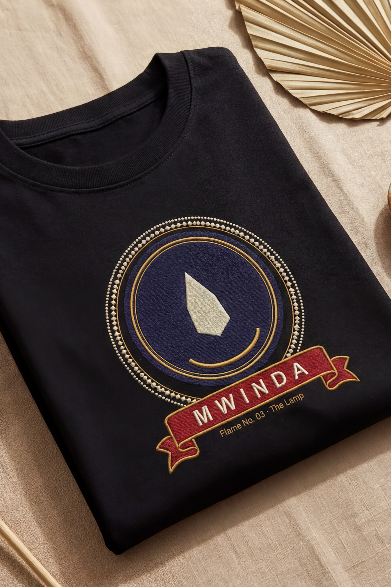 Black Mwinda tee folded flat on natural linen, lamp flame medallion, The Twelve Flames by Bana Bilaka