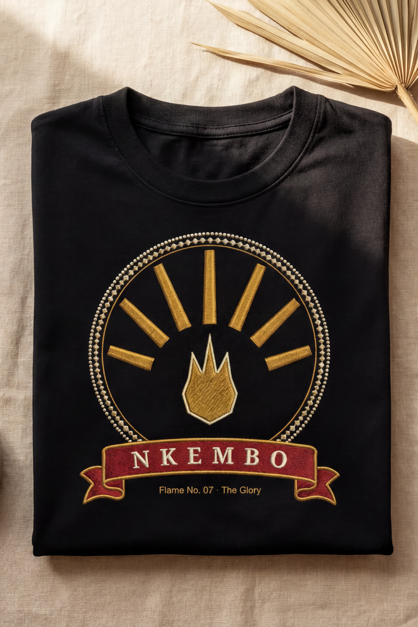 Black Nkembo tee folded flat on natural linen, gold glory medallion, The Twelve Flames by Bana Bilaka
