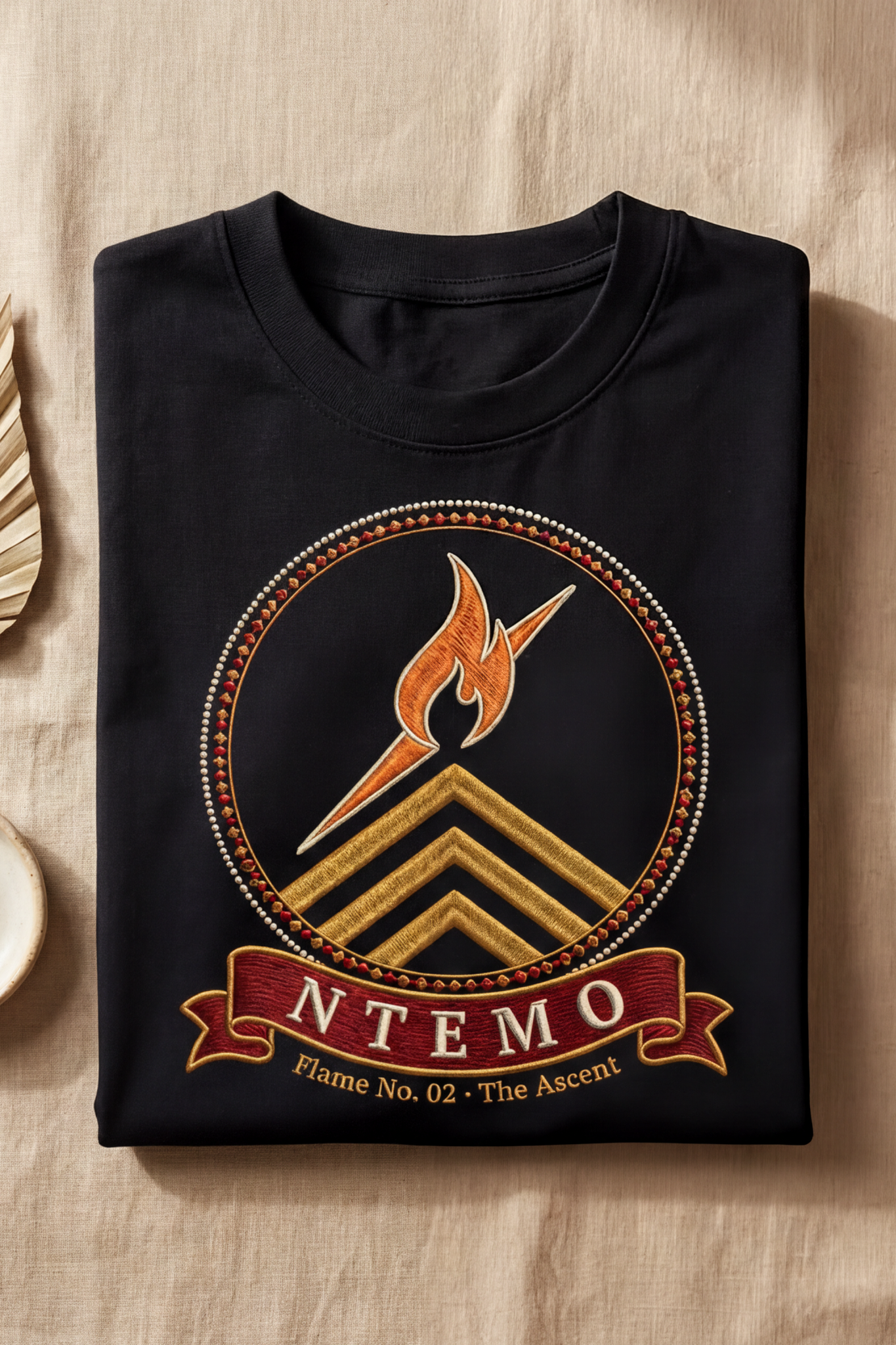 Black Ntemo tee folded flat on natural linen, embroidery-style ascent flame medallion, The Twelve Flames by Bana Bilaka