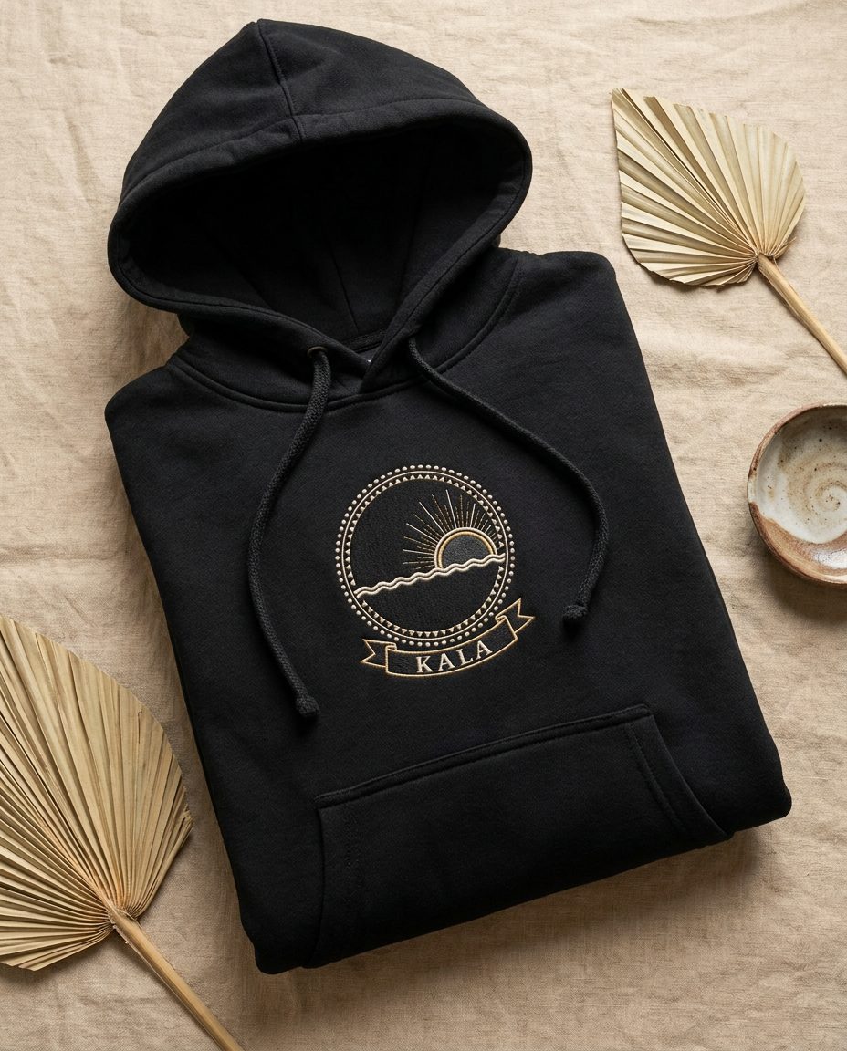 Kala jet black hoodie flat-lay on beige linen, embroidery-style Kongo Dikenga medallion (half-risen dawn sun breaking the Kalunga line on the right) printed on the chest, Bana Bilaka